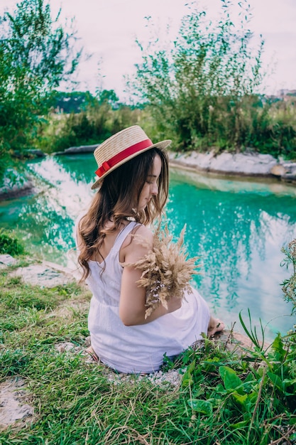 Jovem garota em um vestido branco e um chapéu à beira do lago. mulher sorridente em um vestido com um buquê de flores secas perto de um pequeno lago.