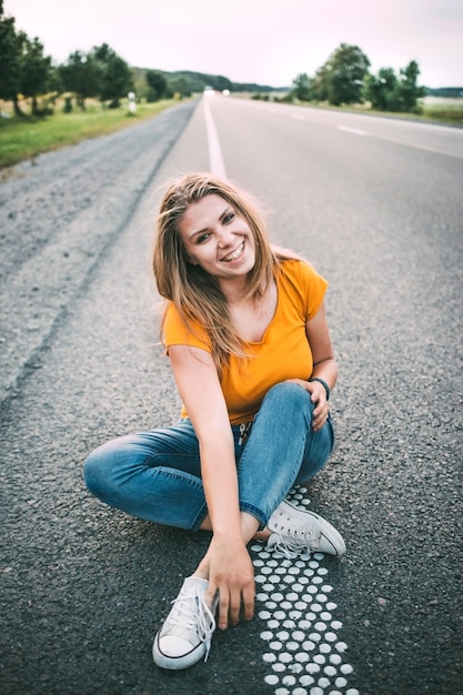 Jovem garota em uma camiseta amarela e calça jeans azul e tênis branco senta-se na estrada e sorri. noite, luz suave.