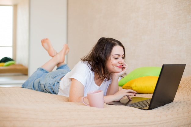 Jovem garota está deitada em casa em uma cama com café e um laptop
