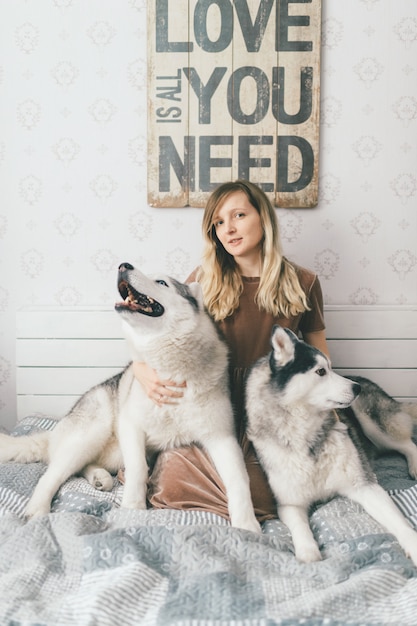 Jovem garota feliz no vestido marrom, sentado na cama e abraços adorável com sorridentes focinhos husky cachorros.