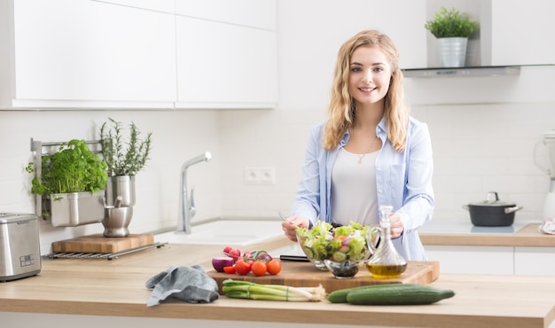 Jovem garota loira feliz preparando salada saudável na cozinha de casa.