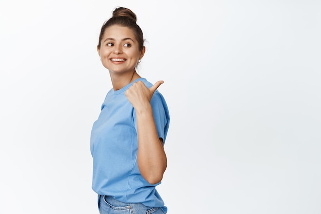 Jovem garota moderna apontando o dedo para a direita e olhando por trás do ombro, mostrando anúncio de logotipo, fundo branco, vestindo camiseta azul