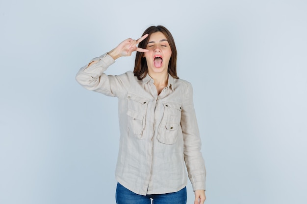 Jovem garota mostrando o sinal de v, mostrando a língua em uma camisa bege, jeans e parecendo se divertindo. vista frontal.