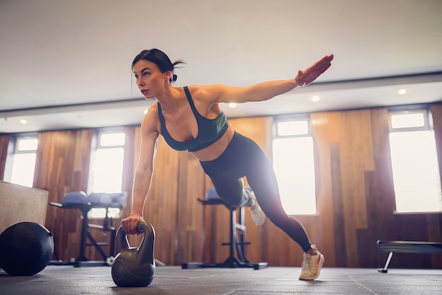 Foto jovem garota motivada fazendo exercícios de prancha usando kettlebells com uma das mãos na academia, foto de corpo inteiro, espaço de cópia