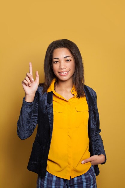 Jovem garota negra apontando para cima e sorrindo em fundo amarelo colorido