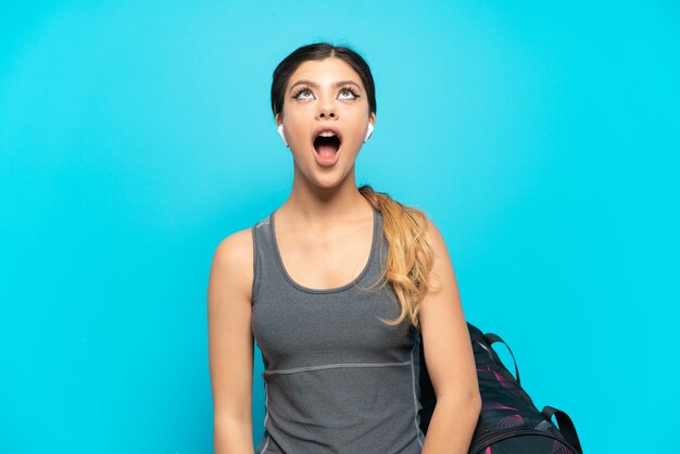 Jovem garota russa com uma bolsa esportiva isolada em um fundo azul, olhando para cima e com expressão de surpresa