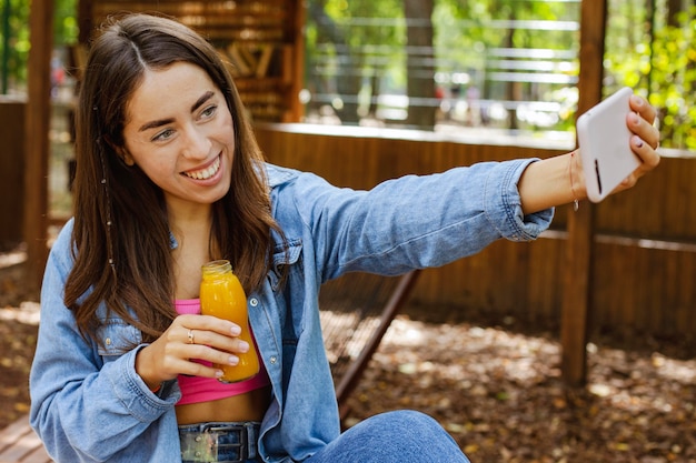 Jovem garota segurando uma garrafa de suco fresco e tirando uma selfie