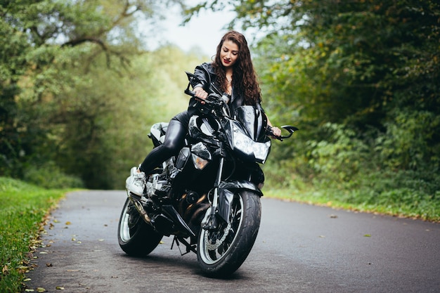 Jovem garota sexy em uma jaqueta de couro preta sentada em uma motocicleta esportiva preta em uma estrada na floresta