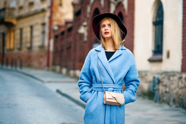 Jovem garota sorridente bonita em um casaco azul e chapéu cor de vinho, andando na rua na sunset