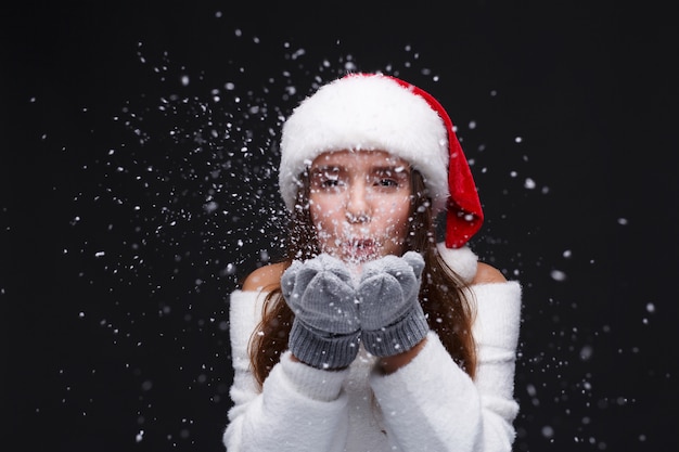 Jovem garota sorridente bonita no chapéu de papai noel vermelho está soprando flocos de neve brancos