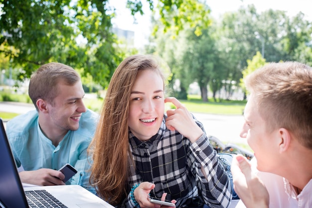 Jovem garota sorridente feliz com dois amigos do sexo masculino relaxando ao ar livre com seus telefones e um laptop, ambiente diurno. Conceito de educação, felicidade e amizade.