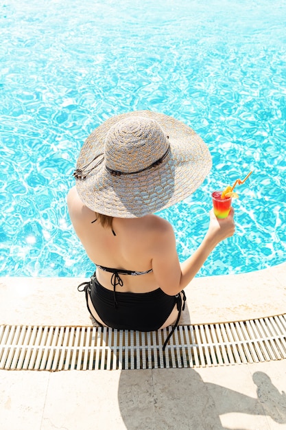 Jovem garota usando chapéu de sol localização junto à piscina com cocktail