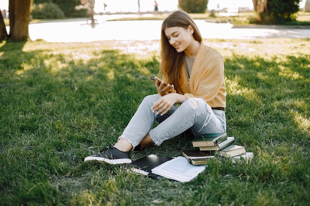 Jovem garota usando um smartphone enquanto está sentado em uma grama. Mulher tem livros perto dela