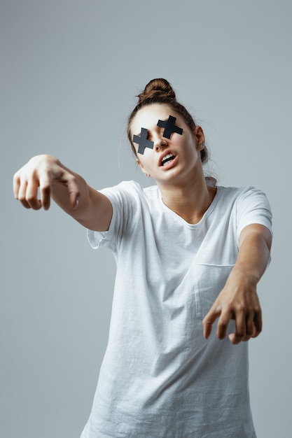 Jovem garota vestida de camiseta branca e jeans com cruzes pretas de fita adesiva nos olhos fica no fundo branco no estúdio como um zumbi.