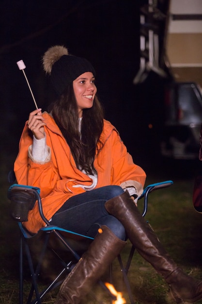 Jovem garota vestindo roupas quentes em uma noite fria de outono em um acampamento assando marshmallow. Van de campista retrô em segundo plano.
