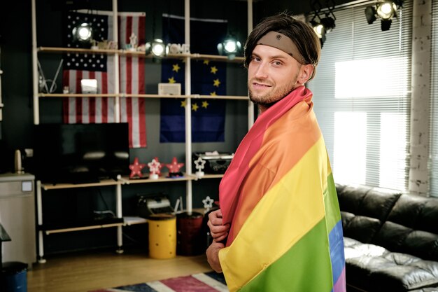 Foto jovem gay com bandeira do orgulho em pé na sala e olhando para a câmera