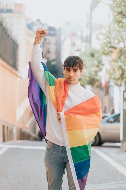 Jovem gay envolto em uma bandeira do arco-íris em pé com o punho erguido no meio da rua. conceito de comunidade lgbt, direitos das minorias, proteção dos direitos humanos. cidade de madrid