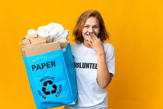 Jovem georgiana segurando uma sacola de reciclagem cheia de papel para reciclar feliz e sorridente, cobrindo a boca com a mão