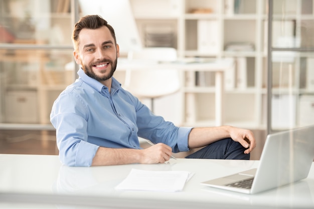 Jovem gerente alegre com um sorriso largo, sentado à mesa no escritório com o laptop na frente e trabalhando no planejamento