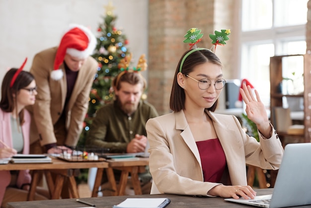 Jovem gerente asiática sorridente com fita de natal olhando para o visor do laptop e acenando com a mão enquanto cumprimenta o cliente ou colega