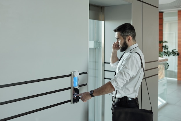 Foto jovem gerente de escritório ocupado com smartphone falando com o cliente enquanto está perto do elevador e aperta o botão de chamada