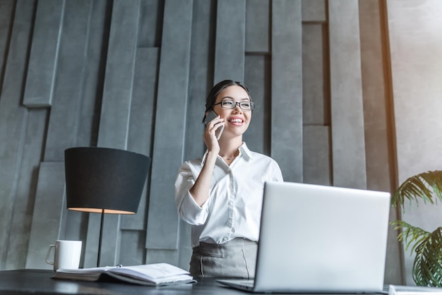 Jovem gerente feminina consultoria no telefone e usando laptop no local de trabalho