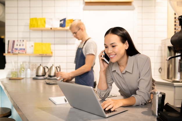 Jovem gerente positiva de um restaurante que consulta clientes ao telefone em frente ao laptop enquanto sua colega prepara café para os convidados