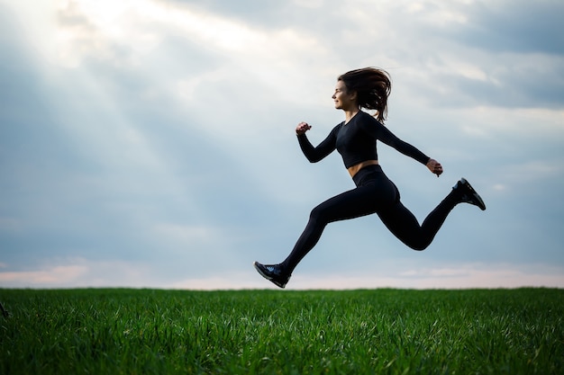 Jovem ginasta profissional está pulando na natureza contra o céu azul. atleta garota em um top preto e legging preta fazendo exercícios acrobáticos