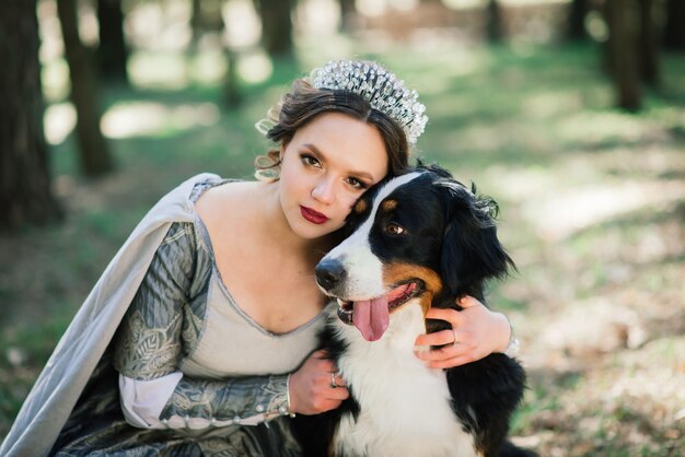 Jovem glamourosa linda senhora usando vestido luxuoso e casaco em pé na floresta com seu cachorro.