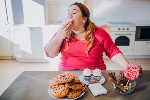 Foto jovem gorda na cozinha, sentado e comendo alimentos doces. gula.