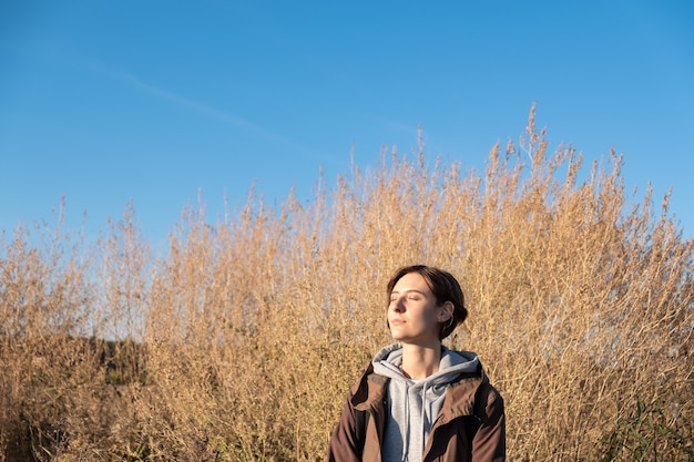 Jovem gosta do sol de outono. Mulher em parka ao ar livre em um prado em uma tarde ensolarada