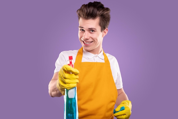 Jovem governanta sorridente, confiante, com um avental amarelo e luvas, demonstrando uma garrafa de plástico com detergente de limpeza contra um fundo violeta