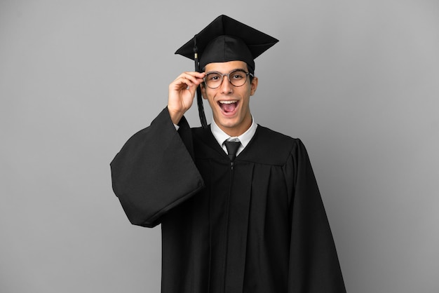 Jovem graduado universitário argentino isolado em um fundo cinza com óculos e surpreso