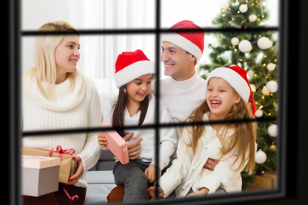 Jovem grande família celebrando o Natal desfrutando do jantar, vista de fora através de uma janela para uma sala de estar decorada com luzes de árvore e velas, pais felizes comendo com três filhos.