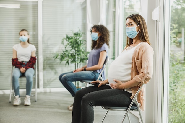 Jovem grávida com uma máscara facial sentada em uma sala de espera e esperar o check-up do ginecologista.