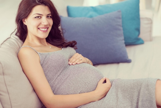 jovem grávida feliz mantendo a mão na barriga enquanto está sentada no sofá em casa