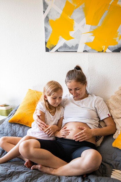 Foto jovem grávida feliz sentada no sofá com a filha acariciando a barriga
