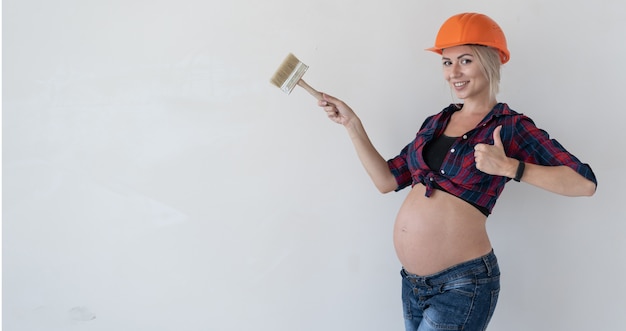 Jovem grávida fica em um fundo branco. Vestido com roupas de trabalho. Camisa xadrez vermelha e capacete de construção. Segure o pincel em suas mãos. Indica um espaço vazio e levanta o polegar.