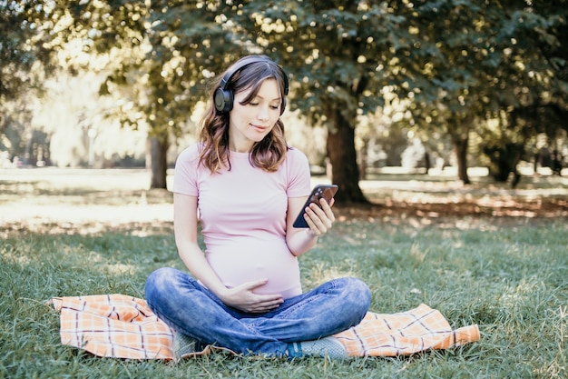 jovem grávida ouvindo música e usando seu telefone no parque