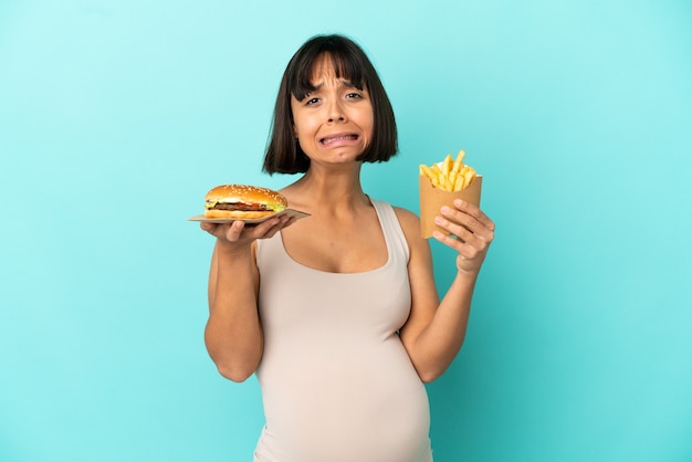 Jovem grávida segurando hambúrguer e batatas fritas sobre fundo azul isolado