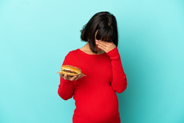 Jovem grávida segurando um hambúrguer sobre uma parede isolada com uma expressão cansada e doente