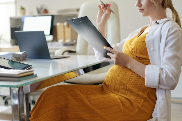 Foto jovem grávida sentada à mesa no serviço social e preenchendo documentos