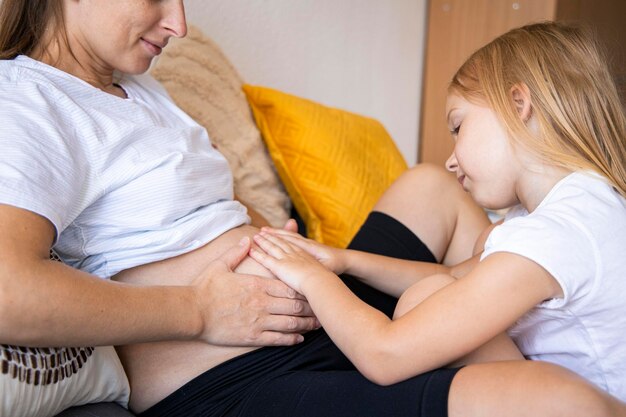 Foto jovem grávida sentada no sofá com a filha acariciando a barriga