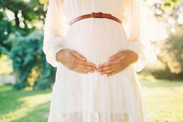 Jovem grávida vestida de branco com as mãos sobre a barriga no fundo verde da natureza