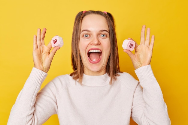 Jovem gritando muito feliz com rabos de cavalo em suéter mostrando marshmallow delicioso e açucarado posando isolado sobre fundo amarelo gritando de excitação quebra dieta