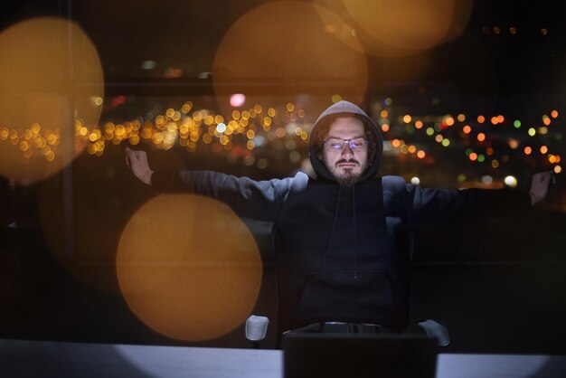 Jovem hacker talentoso usando laptop enquanto trabalhava em escritório escuro com luzes da cidade grande ao fundo à noite