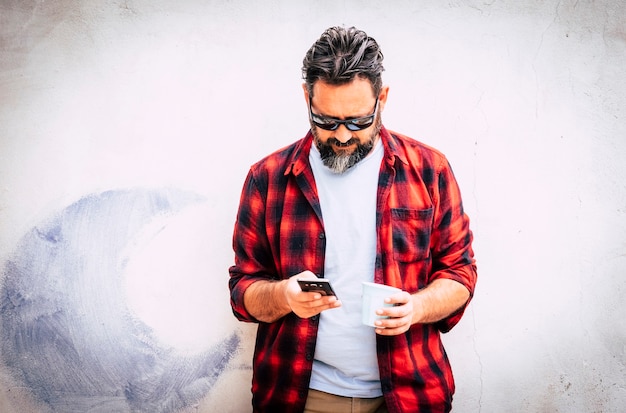 Jovem hippie caucasiano com barba e camisa vermelha olhando para o telefone para mensagens ou ligação de amigos