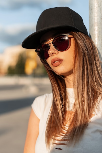 Jovem hippie de retrato de beleza com lábios sensuais em branco top rasgado em óculos de sol da moda em elegante boné de beisebol legal no sol ao ar livre. mulher bonita e moderna em roupas jovens muamba na cidade ao pôr do sol.