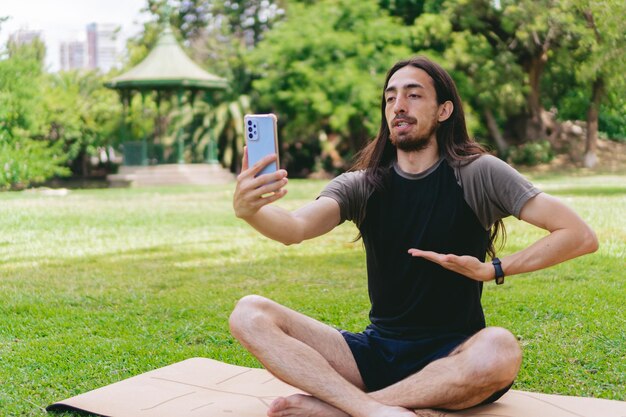 Jovem hippie latino e hispânico com cabelo comprido e barba inalando enquanto participava de uma videochamada de um campo verde sentado em um tapete de ioga em posição de lótus