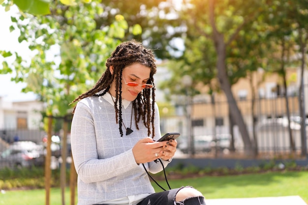 Jovem hipster com dreadlocks em óculos de sol rosa usa smartphone sentado no parque de verão ao ar livre Nova geração Tecnologia na mão Verão